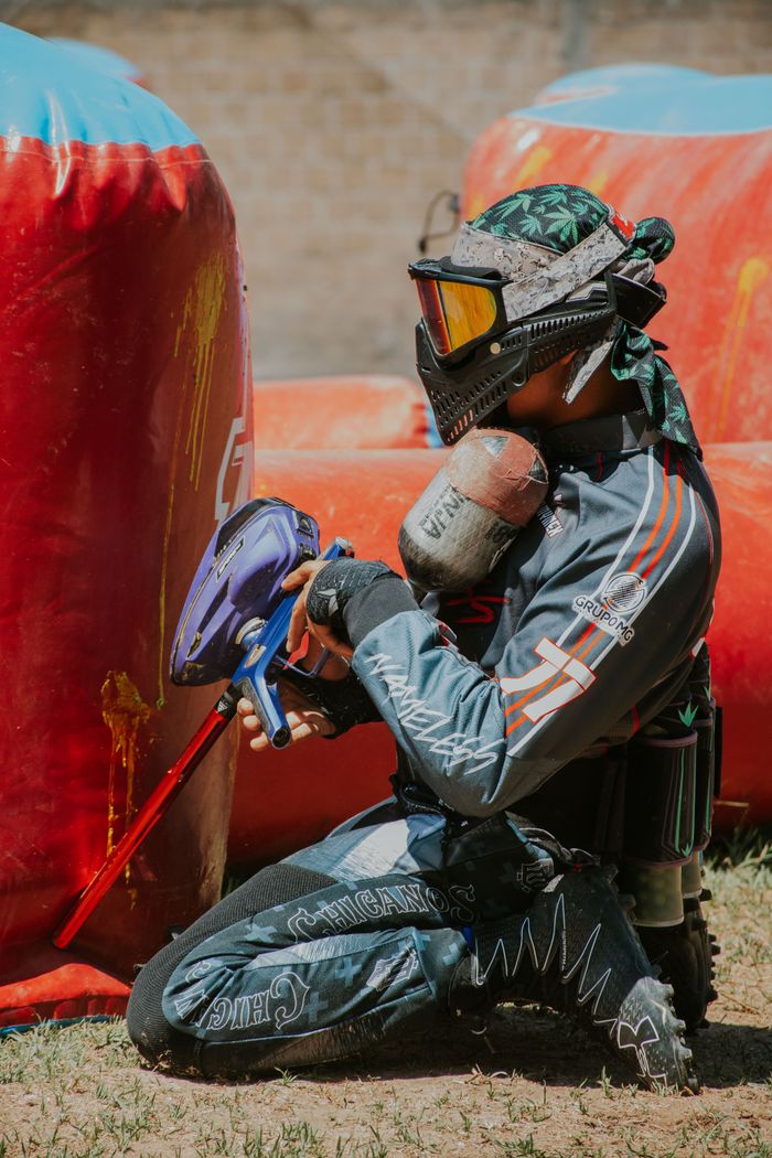 services-02 A paintball player kneels and aims, fully equipped with mask and gear in a competitive match outdoors.