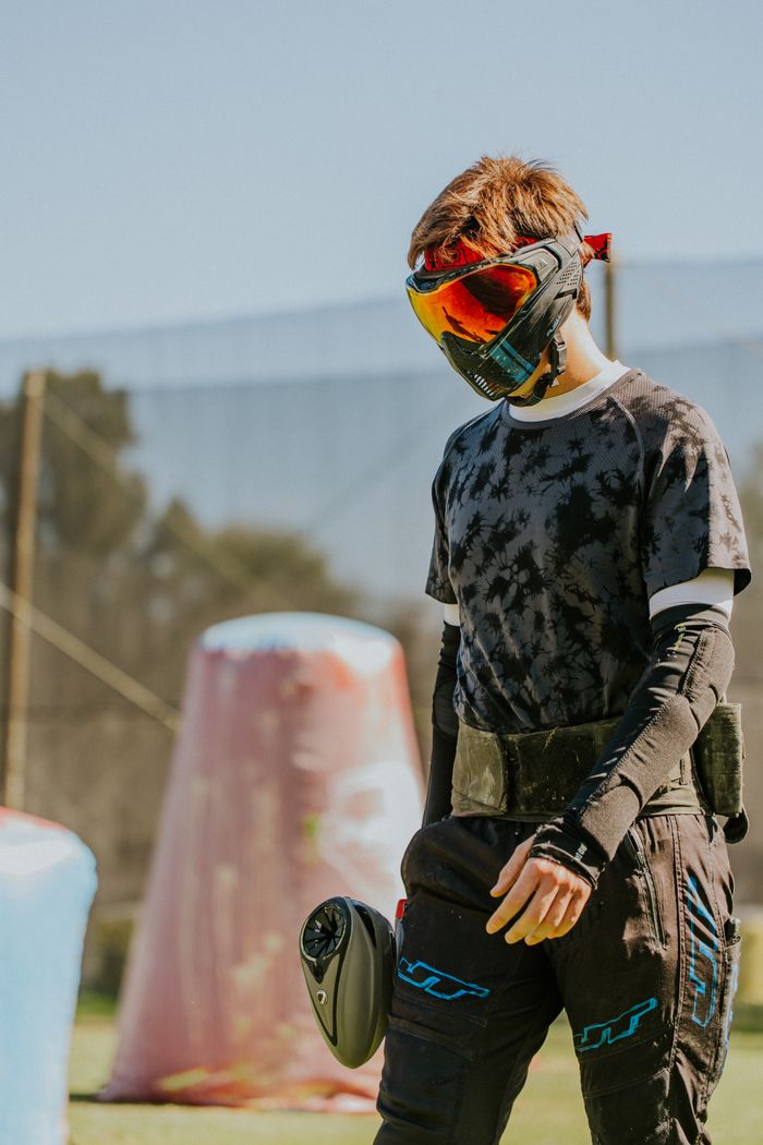 Teenager wearing paintball protective gear at an outdoor paintball field on a sunny day.