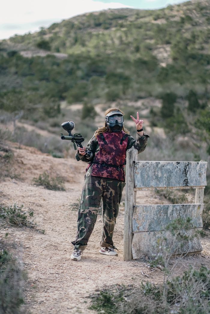 services-01 A woman in camouflage gear and mask stands outdoors with a paintball gun, making a V-sign.